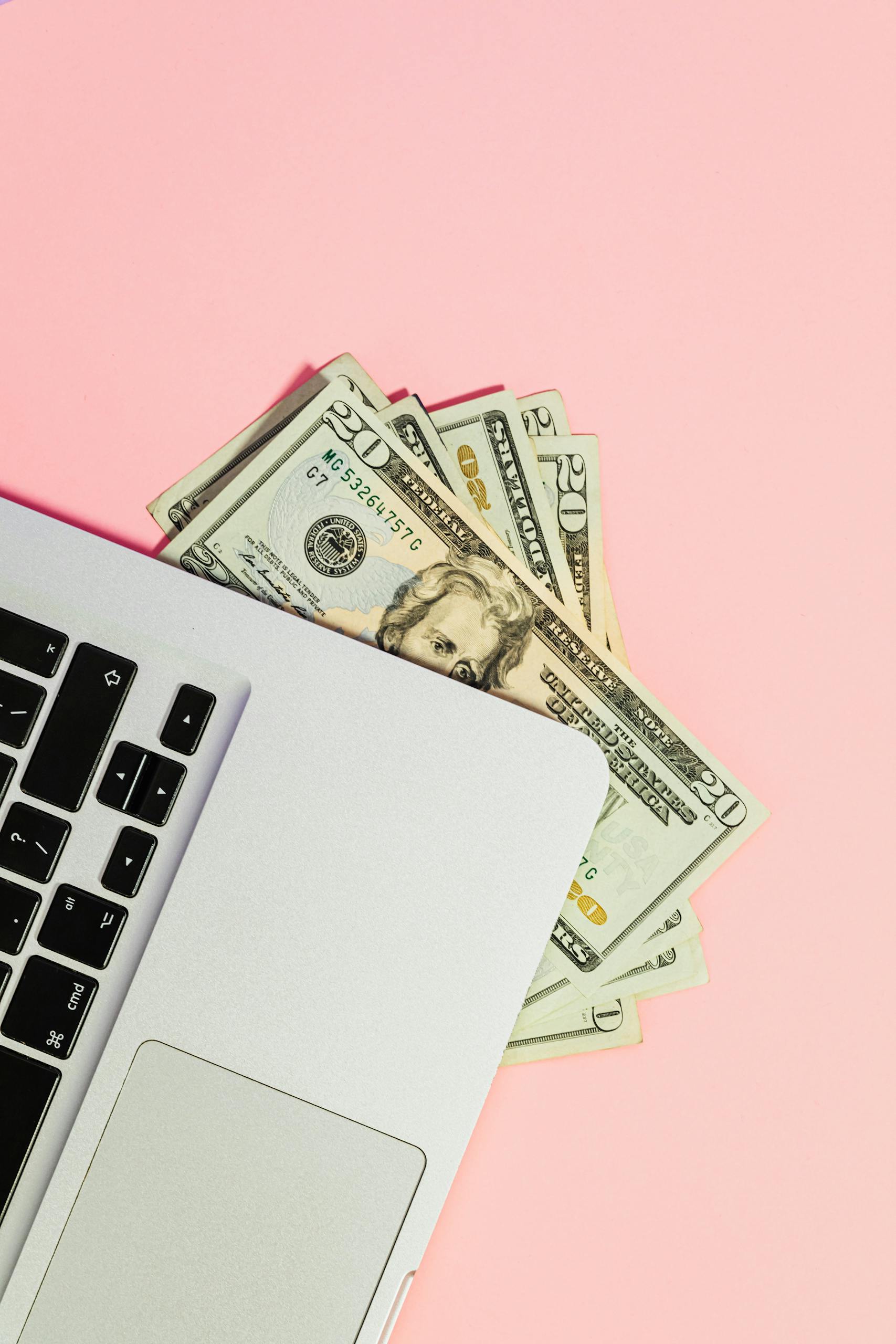 Overhead shot of dollar bills tucked under a laptop on a pink background, symbolizing online transactions.